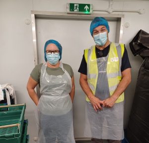 two people dressed in protective aprons, hair and face masks smiling in a catering kitchen.